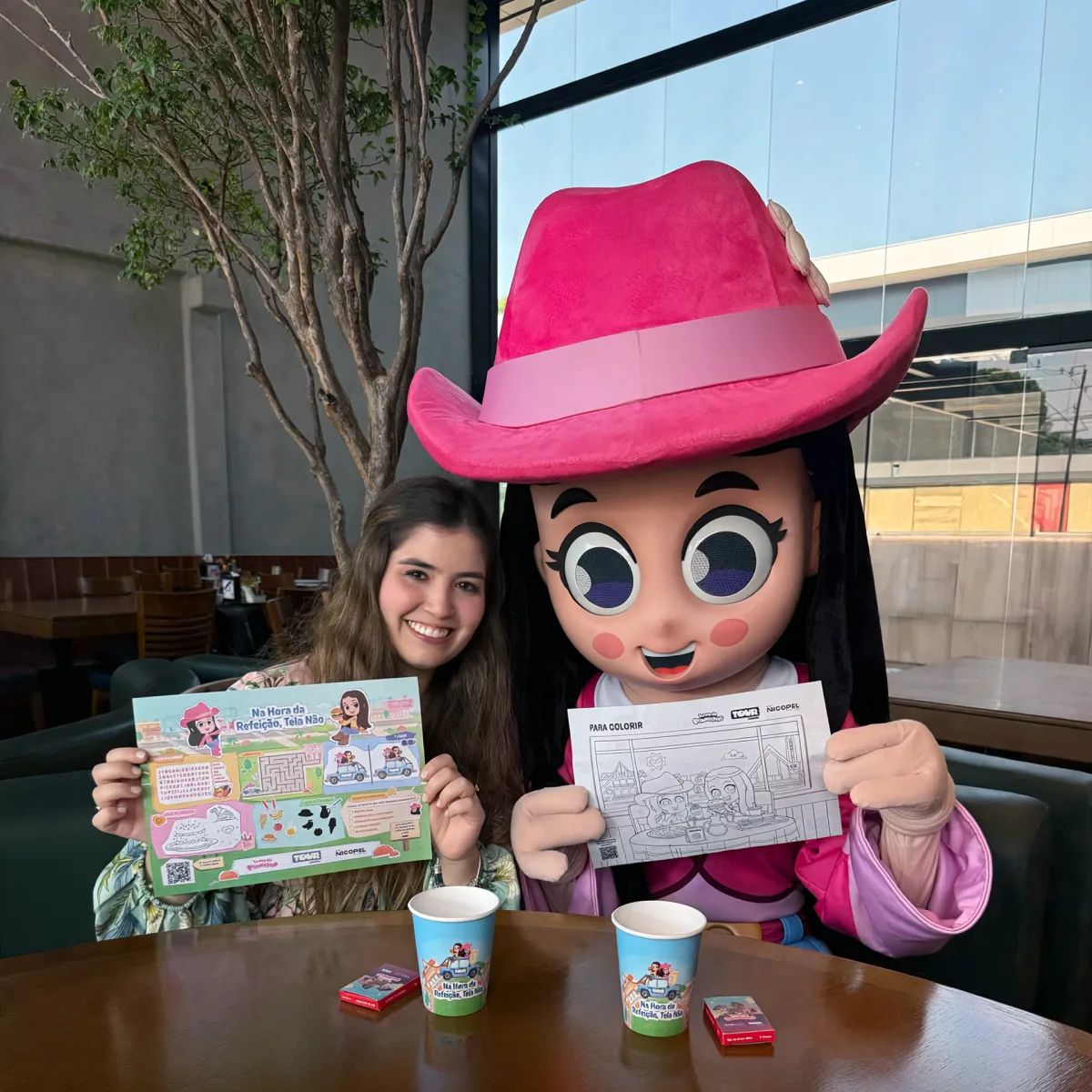 A Boiadeirinha e uma convidada sentadas à mesa de um restaurante, mostrando o papel-bandeja preenchido e a folha de desenho para colorir